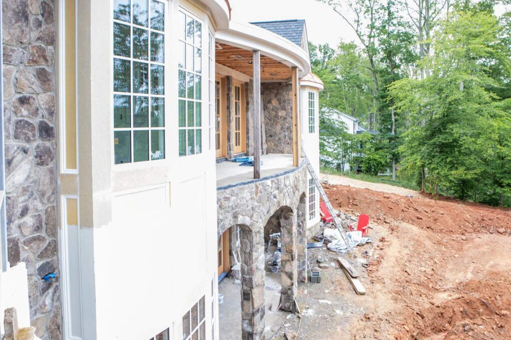 the photo shows an exterior construction phase of a custom home featuring stonework and large windows being installed